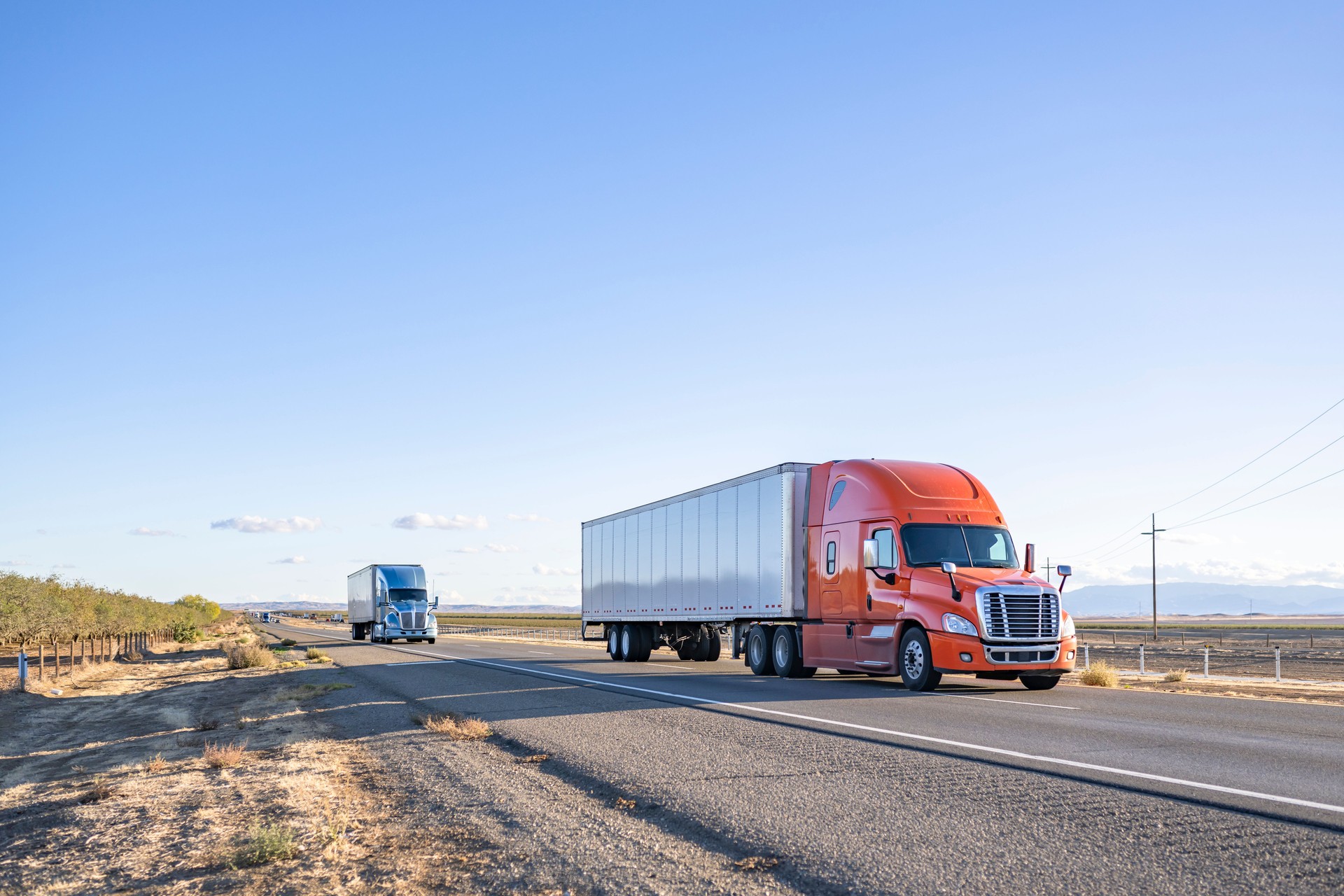 Team of two orange and blue big rigs semi trucks transporting cargo in dry van semi trailers running together on the flat straight highway road in California